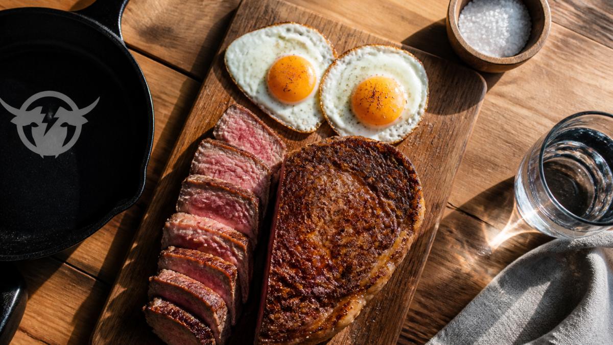 Grass-fed beef steak and fried eggs seasoned with sea salt on a wooden cutting board in morning light