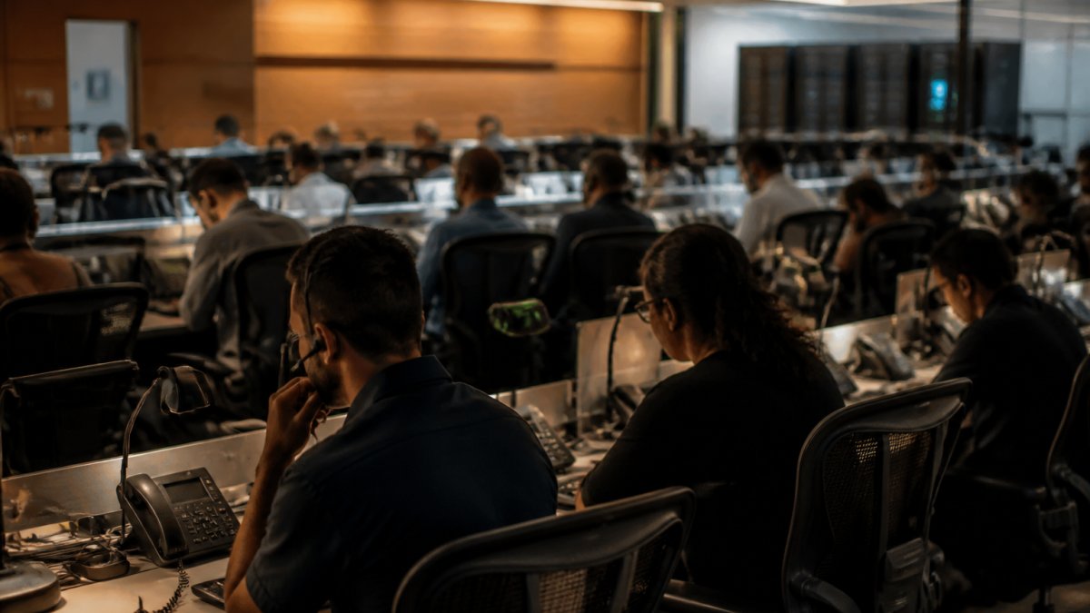IT team members in network operations center monitoring multiple screens displaying real-time system dashboards, alert notifications, and incident response metrics during a critical infrastructure event, with warm overhead lighting illuminating rows of workstations