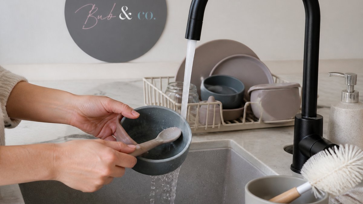 Close-up of a parent's hands washing a silicone baby bowl and spoon under warm running water at a kitchen sink, with a bottle brush and dish rack visible nearby