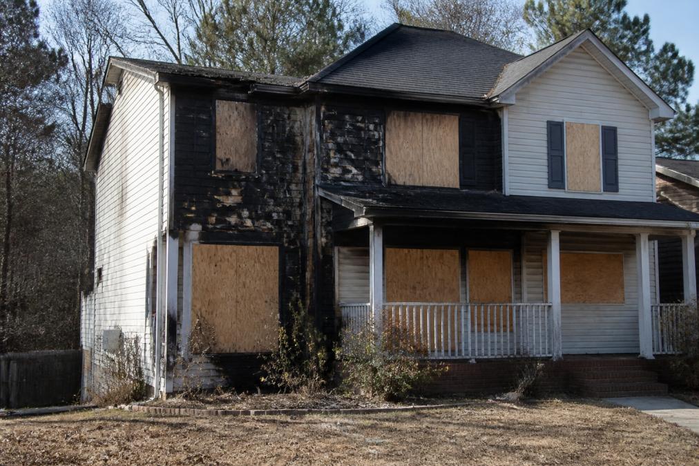 Exterior view of a fire-damaged residential home with boarded-up windows, charred siding visible on one wall, and structural deterioration evident from the street in afternoon lighting