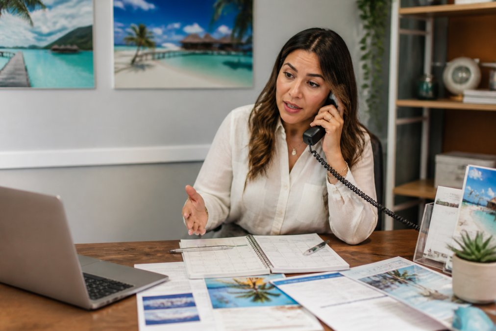 Travel advisor on phone at desk with laptop, calendar, and itinerary documents spread out, supporting a client through flight cancellation situation, office with travel posters on walls