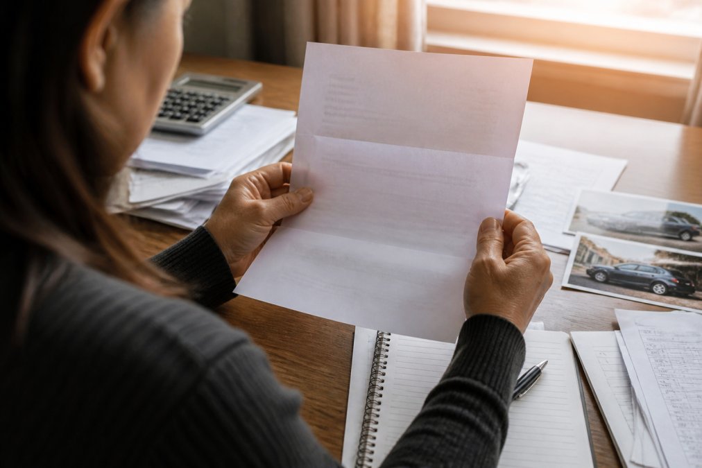 Close-up of a person's hands holding a denial letter at a desk with medical bills, accident photos, and a notepad scattered nearby, natural window lighting