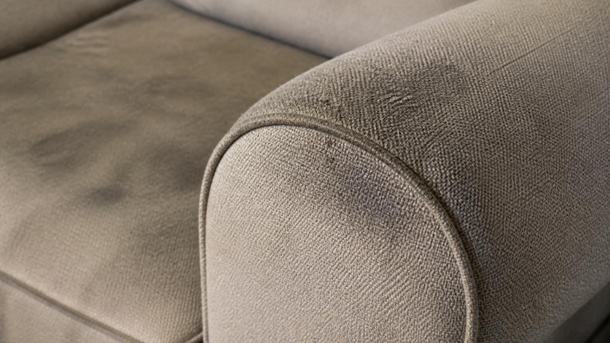 Close-up of upholstered fabric showing visible dust accumulation and darkened areas on armrest and seams with natural light highlighting embedded soil particles