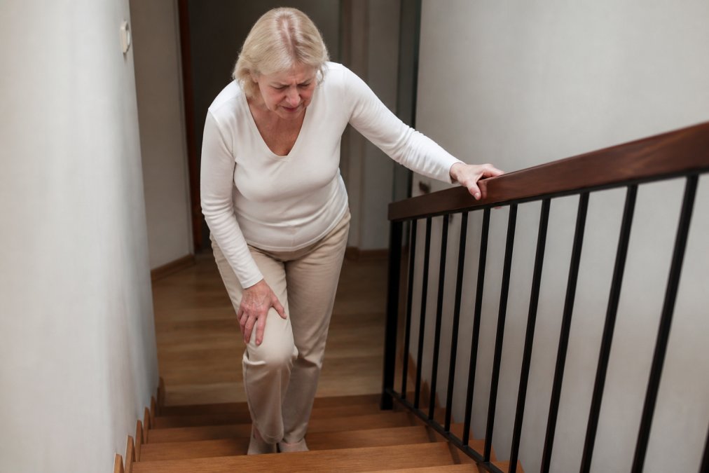 Older adult struggling to climb stairs with visible discomfort, gripping the railing tightly, demonstrating limited range of motion and careful weight distribution