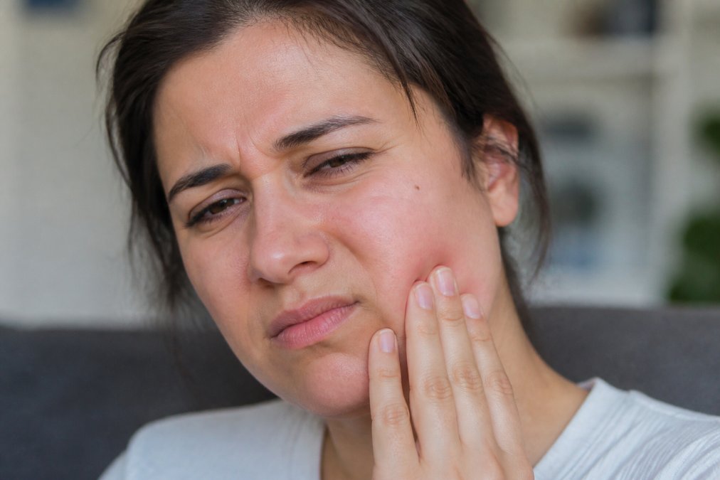 Close-up of a person's face showing visible facial swelling and discomfort, with hand touching the swollen cheek area in soft natural lighting