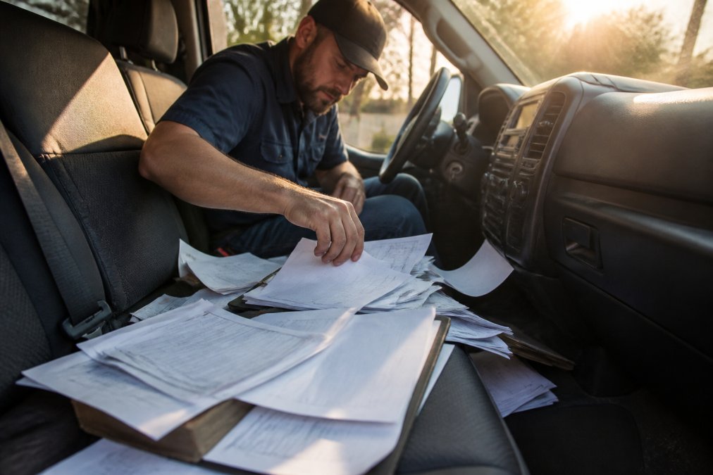 Field technician in truck cab searching through scattered papers and work order documents on seat and dashboard, morning light streaming through windows