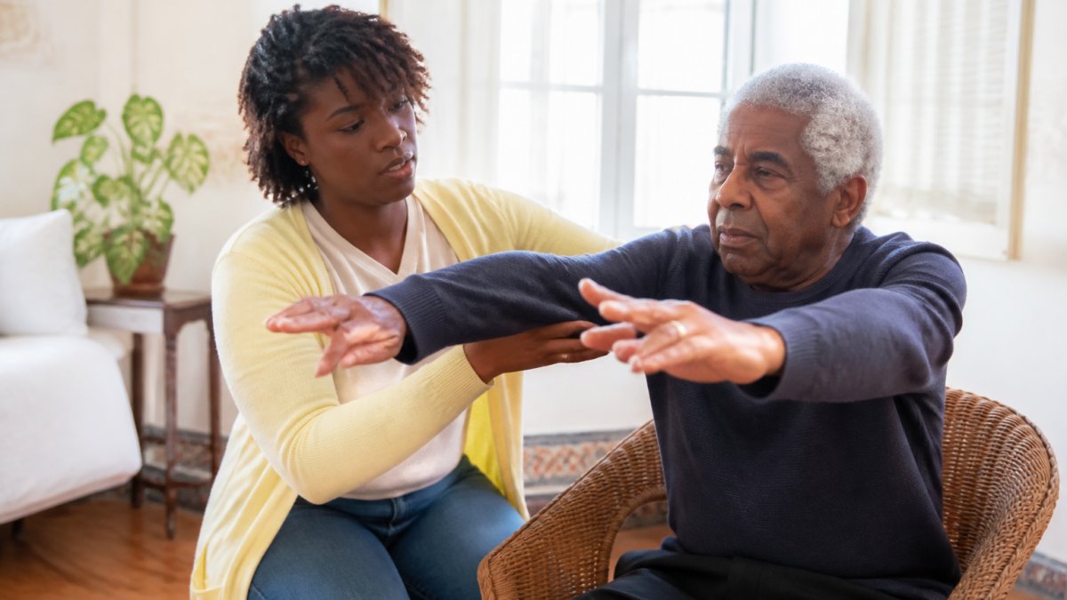 Professional caregiver assisting stroke survivor with guided arm movements and balance exercises in a bright home living room with natural lighting