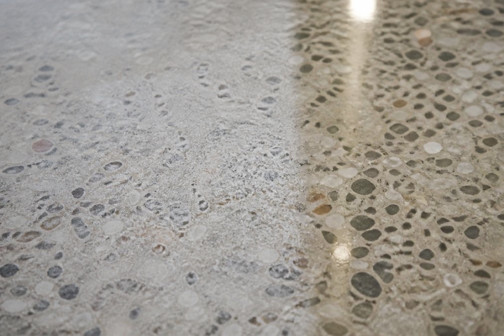 Close-up of terrazzo floor surface showing visible etching marks with dull, chalky appearance and micro-pitting damage contrasted against a nearby polished, glossy section of the same floor