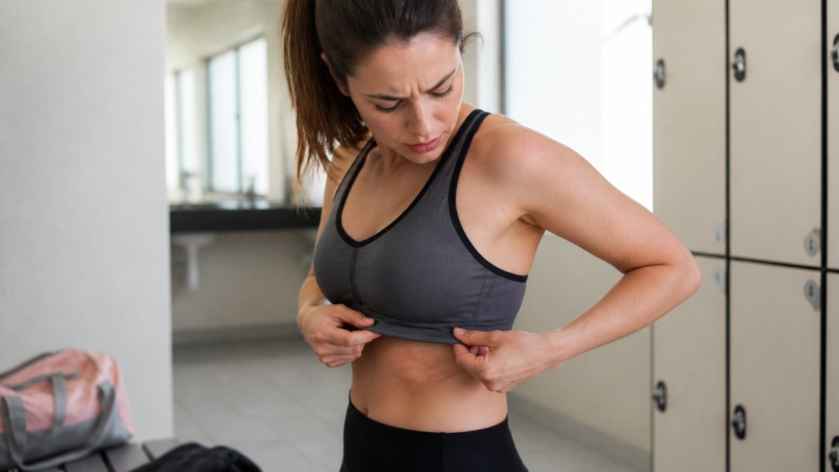 A woman in athletic wear adjusting the band of her sports bra, looking uncomfortable, with visible red pressure marks on her ribcage and shoulders in a bright gym changing room