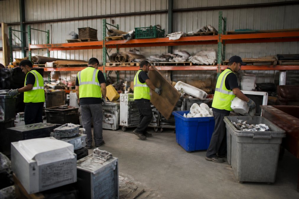 Workers at a sorting facility wearing safety vests organizing furniture, electronics, and household items into separate piles by material type and condition, with metal shelving and organized bins visible in bright industrial lighting