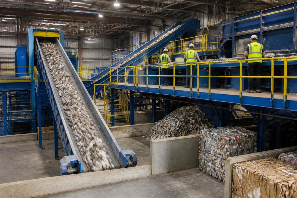 A material recovery facility interior with conveyor belts processing sorted recyclable materials, workers in safety vests overseeing the operation, bright industrial lighting illuminating organized piles of metals, plastics, and cardboard