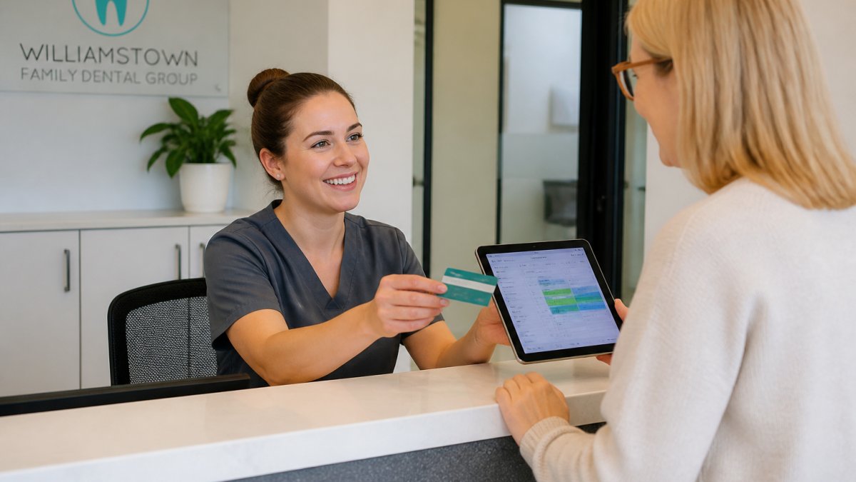 A friendly dental receptionist helping a patient at a bright front desk, holding a health fund card and a tablet showing appointment details