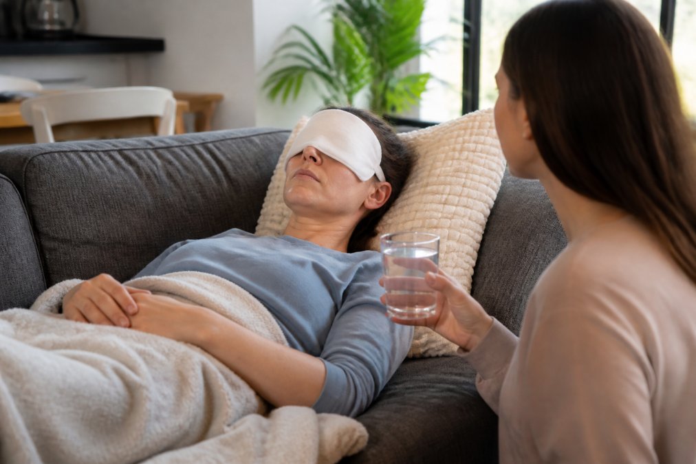 A patient resting on a couch with eyes closed and a cold compress over their eyes, a loved one nearby holding a glass of water, soft natural light in a comfortable living room setting