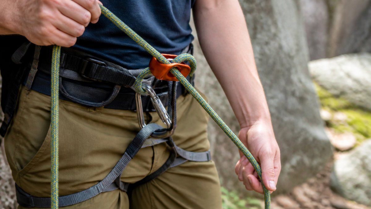 Close-up of a belayer's hands feeding a climbing rope through a belay device attached to a harness belay loop, with the brake strand angled down and the carabiner clearly visible