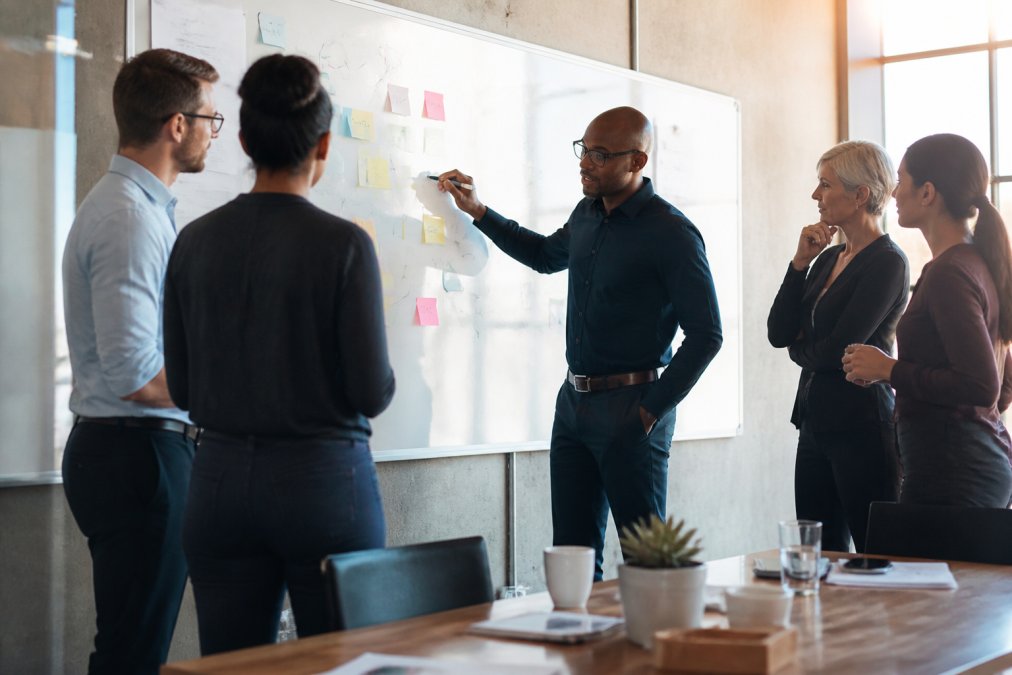 A diverse team of business professionals collaborating around a whiteboard covered with strategy notes, arrows, and sticky notes in a bright modern office