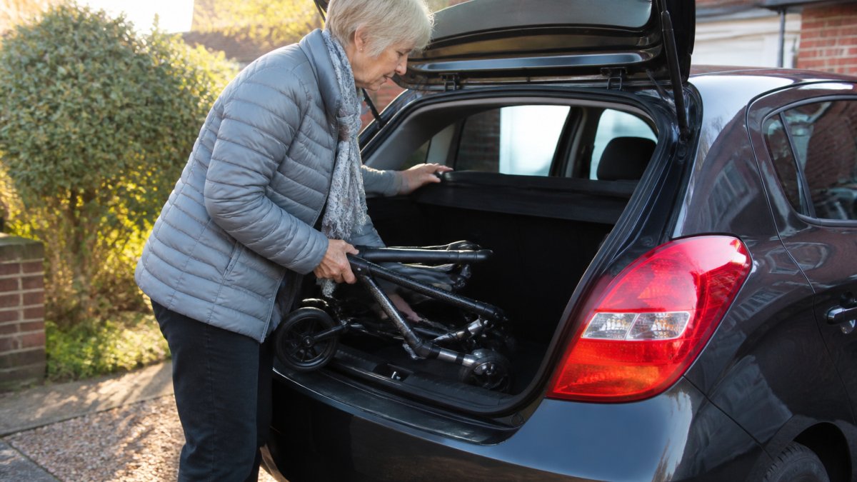 Older woman lifting a lightweight folded rollator into the trunk of a car on a sunny driveway