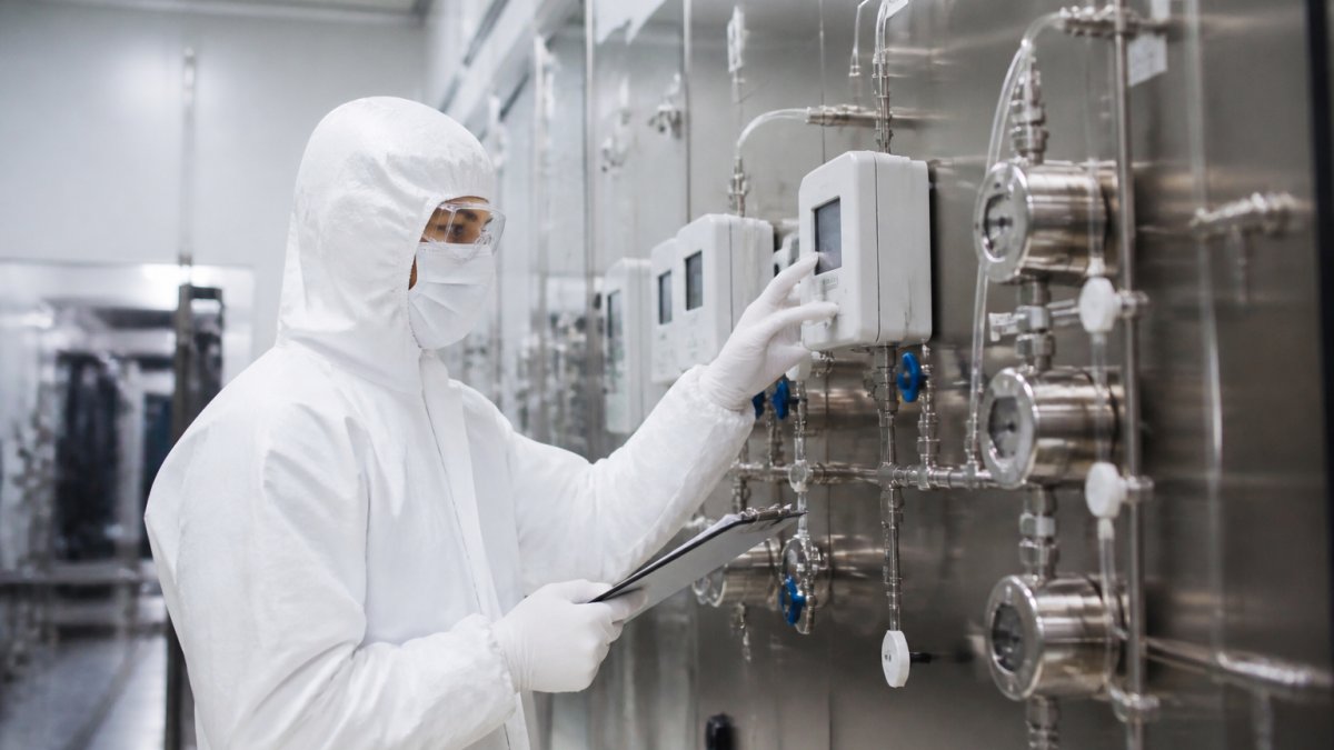 Professional cleanroom technician in full protective gear monitoring environmental sensors and particle counters on a control panel in a modern pharmaceutical facility under bright laboratory lighting