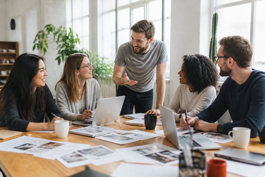 A marketing team gathered around a large monitor reviewing campaign performance dashboards in a bright modern studio office, natural light streaming through windows