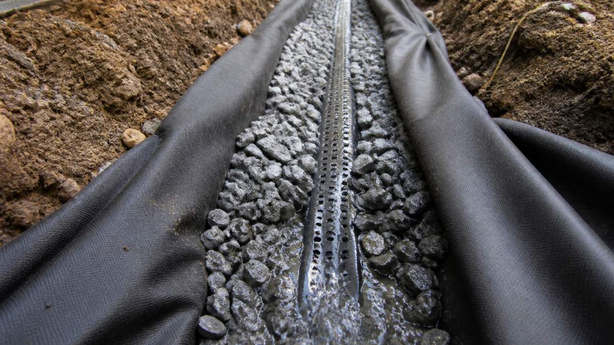 Close-up of exposed trench showing perforated drain pipe surrounded by gray gravel and landscape fabric, with water flowing through the system during installation