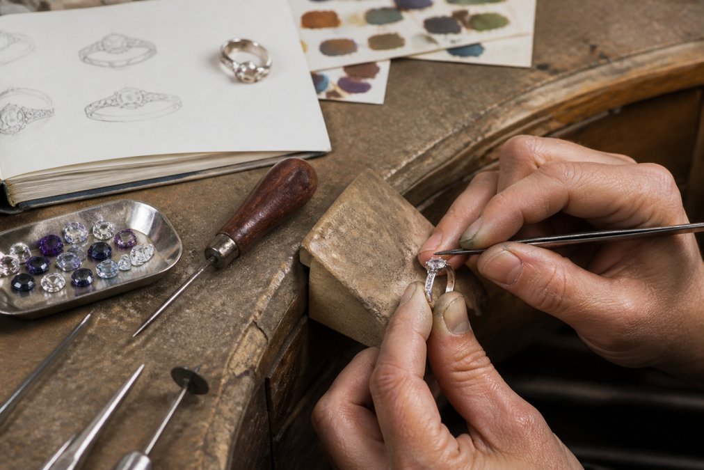 Professional jeweler's hands working on a custom ring design at a workbench, carefully setting a gemstone with precision tools, with sketches and color swatches visible nearby