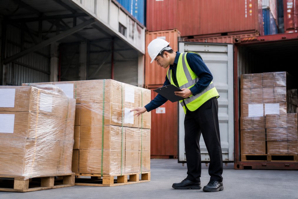 Warehouse worker organizing shipping containers and pallets with freight labels in modern logistics facility, organized cargo visible in background