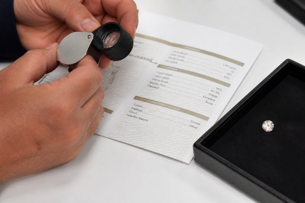 Hands using a jeweler's loupe to inspect a loose diamond and GIA diamond certificate report on a table