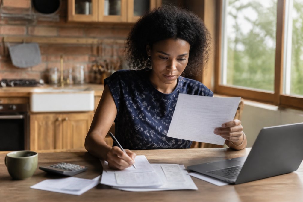 A person sitting at a kitchen table reviewing financial documents and a laptop showing a credit monitoring dashboard, looking focused and determined in warm natural light from a nearby window