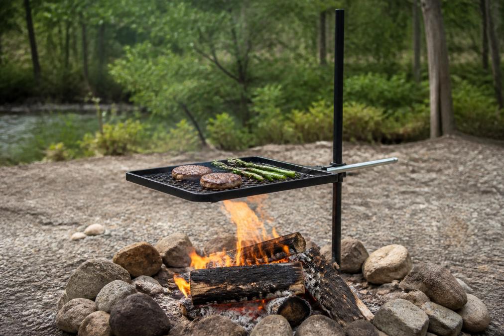 Outdoor campfire cooking scene with a heavy-duty adjustable grill in use over open flames, showing the durability and long-term value of quality cookware
