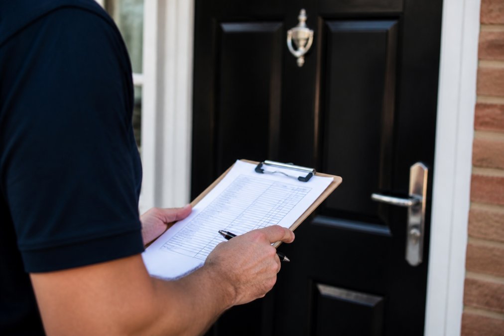 Close-up of a person reviewing an itemised invoice on a clipboard, pen in hand, with a residential front door visible in the background