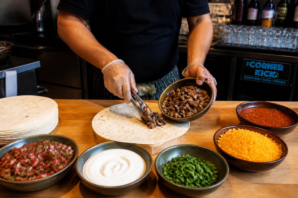 Close-up overhead shot of fresh burrito ingredients arranged on a wooden counter: flour tortilla, carne asada, pico de gallo, sour cream, cilantro, cheddar cheese, and salsa in small bowls