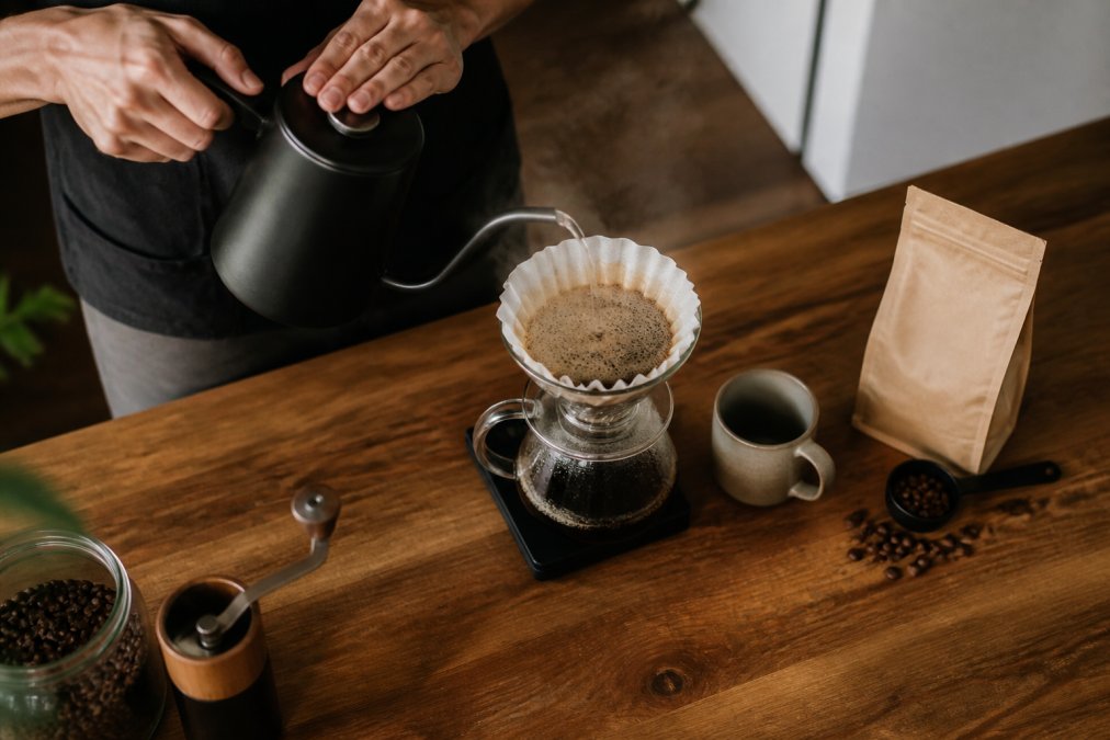 Hands pouring hot water from a gooseneck kettle for a slow coffee ritual over a pour-over dripper on a wooden counter