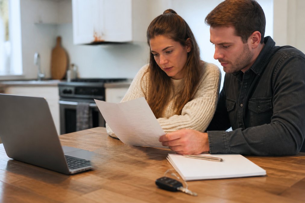 Buyer and seller reviewing a printed vehicle history report at a table with a laptop and car keys