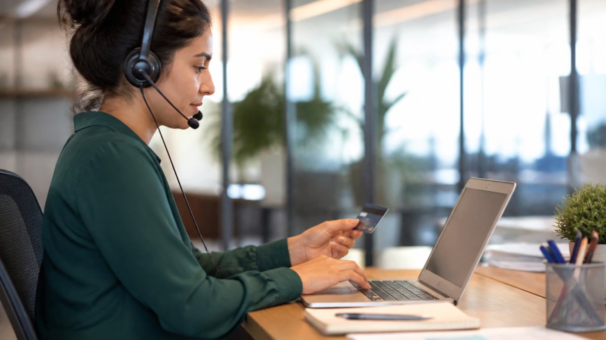 Professional at desk with phone headset entering payment information into a web-based application on computer screen in modern office with natural lighting