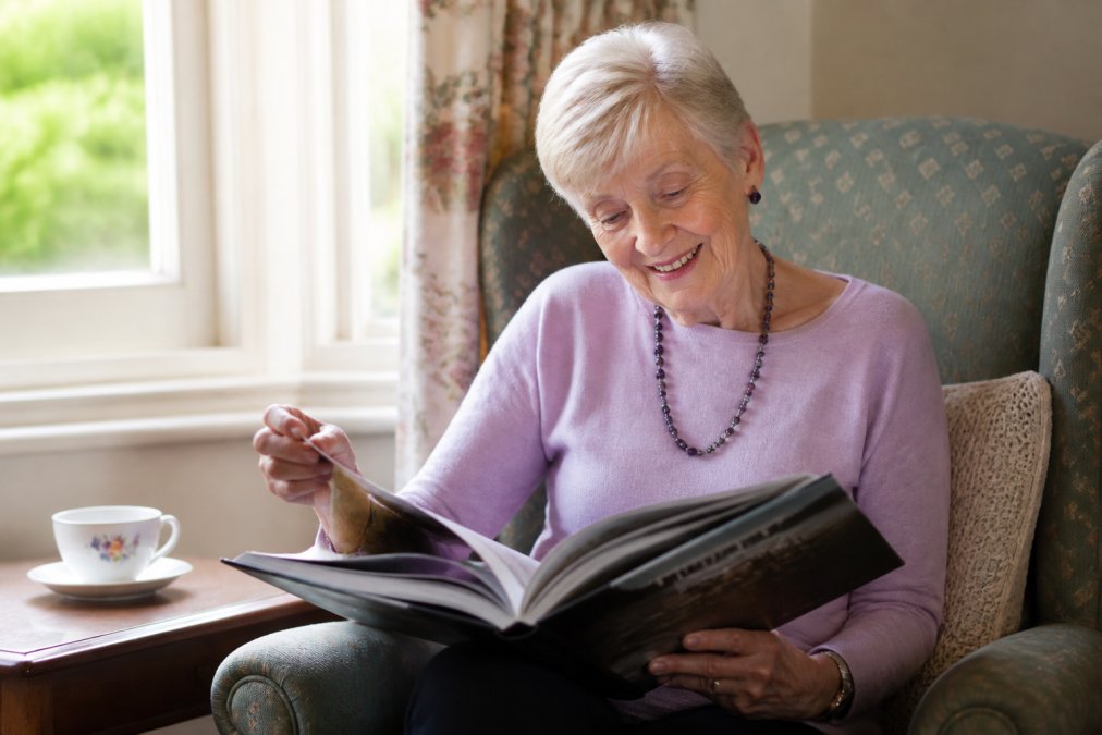 An elderly woman smiling warmly as she turns the pages of a large hardcover nostalgia book, seated in a comfortable armchair near a window with soft natural light, a cup of tea on a side table beside her