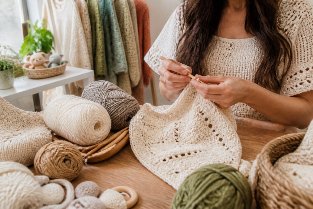 Close-up of hands crocheting a soft natural-fiber garment in a bright home studio, with skeins of yarn and wooden hooks nearby