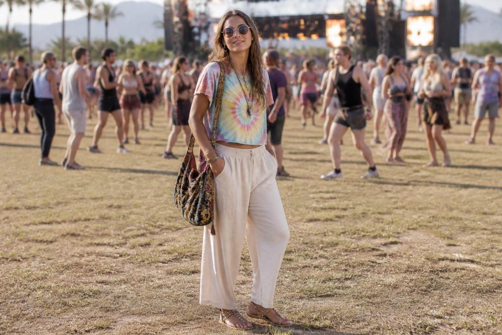 Festival attendee wearing a hand-dyed tie-dye tee, wide-leg trousers in cream linen, embroidered slouchy shoulder bag, and studded sandals, posed in relaxed stance with festival crowd blurred in background