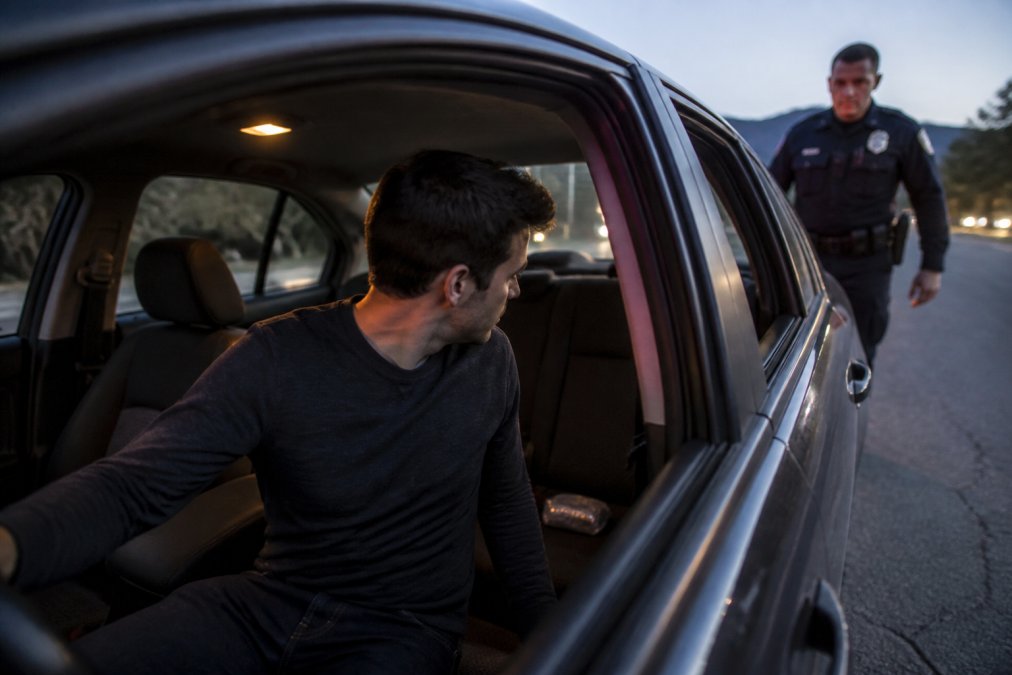 A person sitting in the driver's seat of a car at dusk, looking into the back seat where a small bag is visible on the floor, with a police officer approaching the window