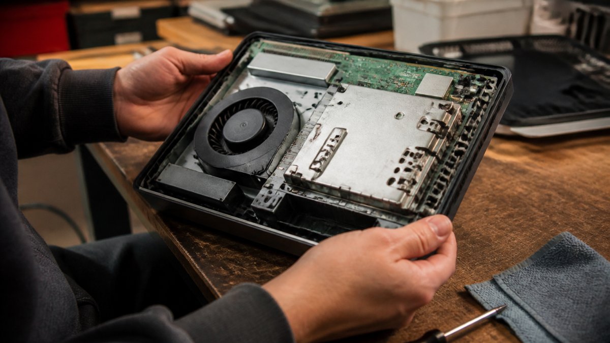 Close-up of hands holding an open gaming console with visible internal components, a screwdriver and cleaning cloth nearby on a workbench, warm workshop lighting