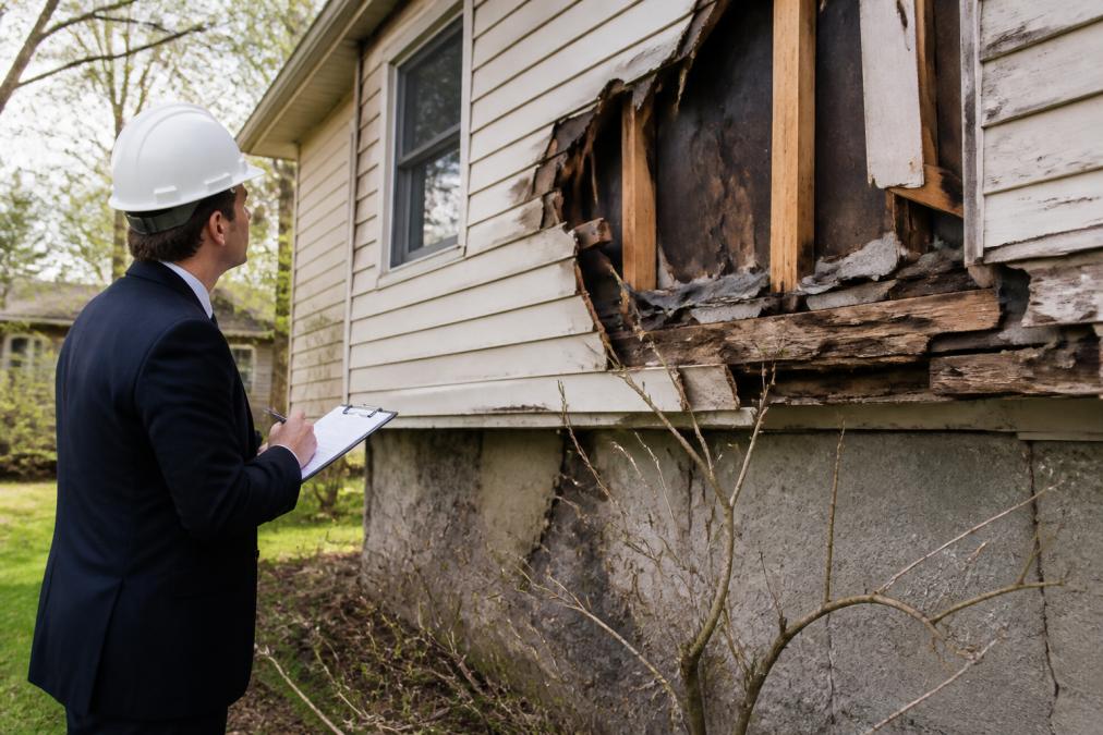 Insurance adjuster in professional attire documenting property damage with clipboard and camera, examining damaged home exterior with visible structural damage in daylight