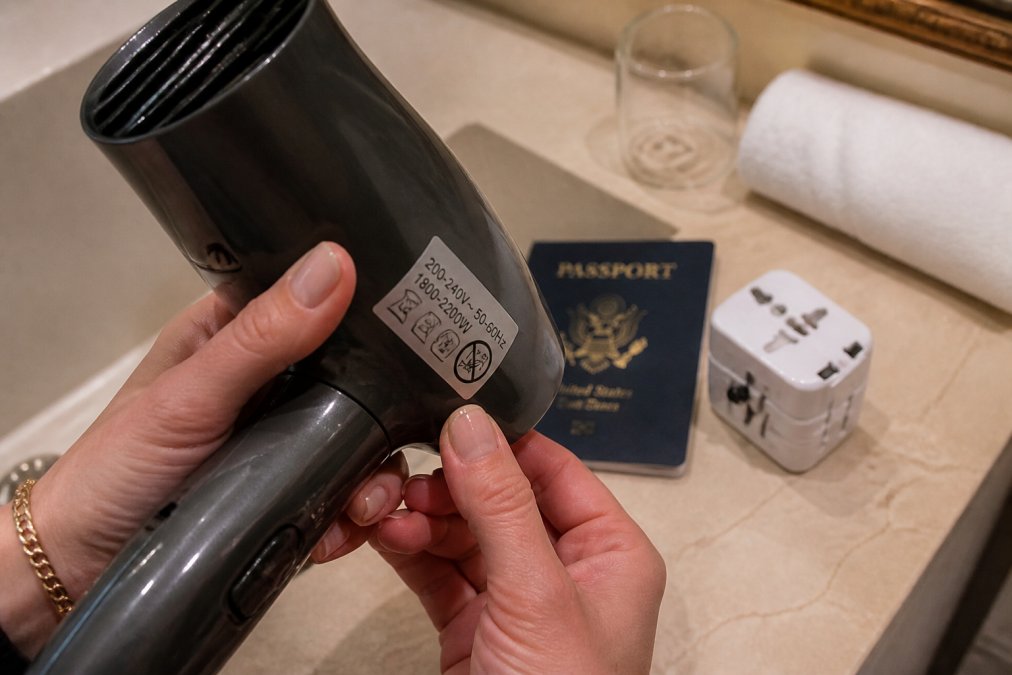 Hands inspecting a hair dryer voltage label next to a passport and travel adapter on a hotel counter