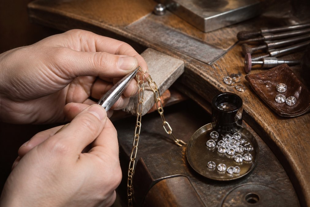 A jeweler's hands using fine pliers to join a hand-made gold chain link on a workbench, with loose diamonds, a loupe and polishing tools arranged nearby in warm studio light