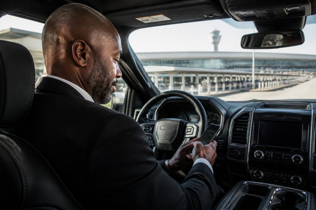 Professional chauffeur in luxury black truck checking flight information on smartphone while parked at Charlotte Douglas International Airport terminal, natural morning light streaming through windshield