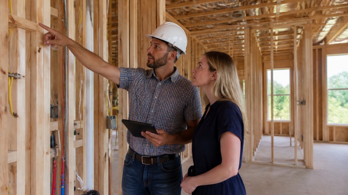 A home inspector and a first-time homebuyer standing inside the unfinished wood framing of a new house, pointing at electrical wiring and plumbing rough-in between wall studs, with natural light coming through open window frames