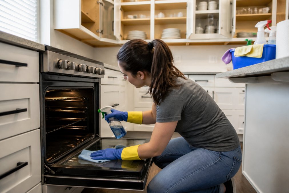 A person in casual clothes cleaning the inside of a kitchen oven with a spray bottle and cloth, wearing gloves, bright kitchen with cabinets open and cleaning supplies on the counter
