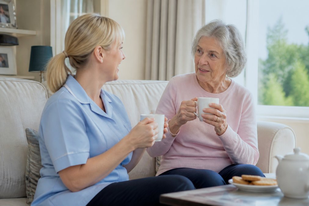 A professional carer in a soft blue uniform sitting beside an older woman on a sofa in a bright living room, sharing a cup of tea and talking warmly