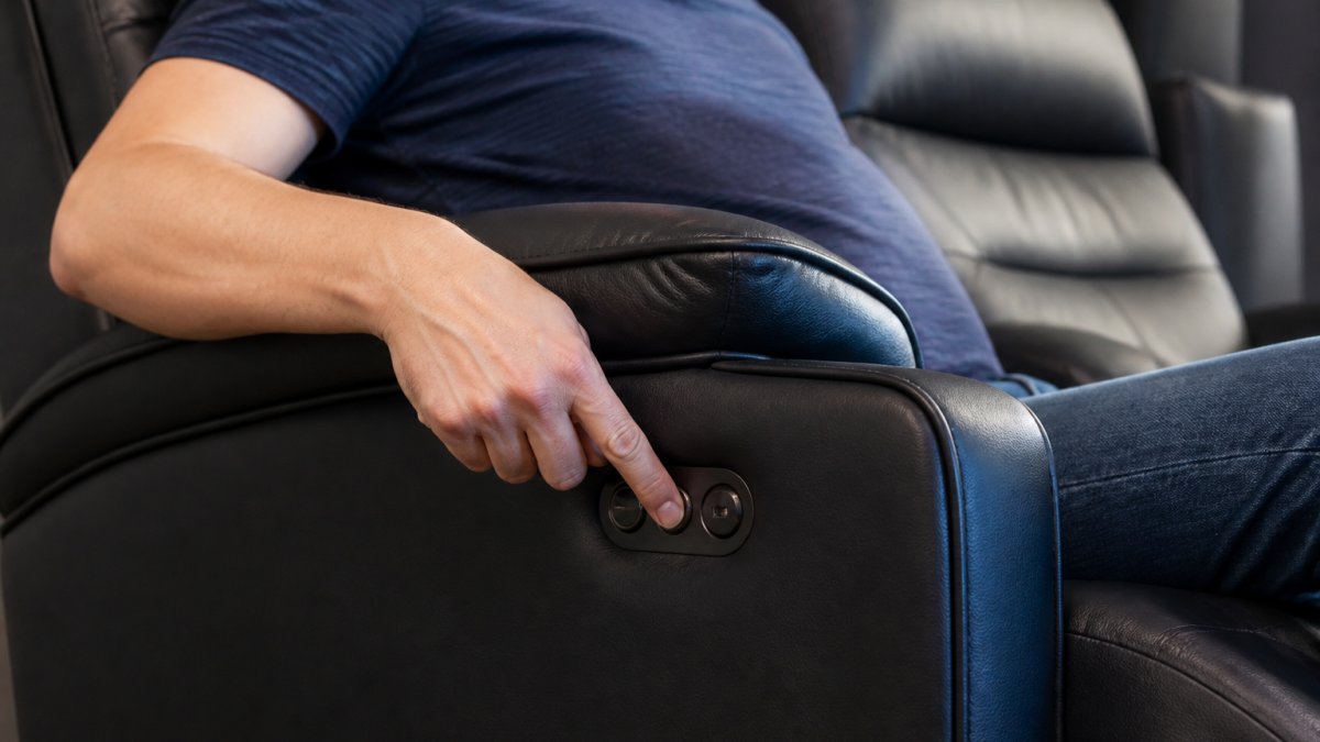 Close-up of a person's hand pressing a power recline button on the side of a black faux leather theater recliner, with the lower back cushion visibly inflated