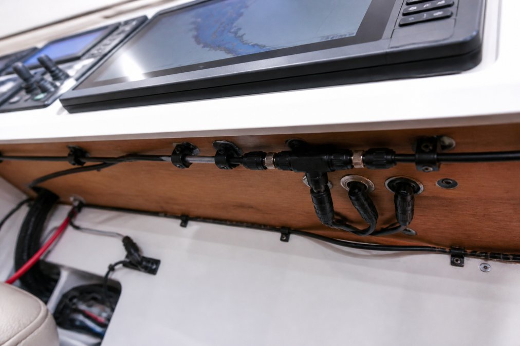 Close-up of a boat helm panel with a Garmin chartplotter, NMEA 2000 backbone cables running along the underside, and a multi-port T-connector joining drop cables, warm cabin lighting reflecting off the instruments
