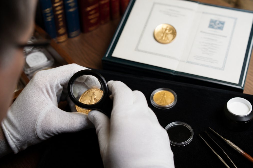 Close-up of hands in white cotton gloves holding a magnifying loupe over a gold coin on a dark felt tray, with a grading certificate visible in the background