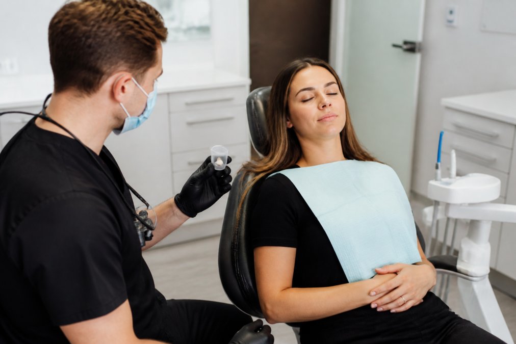 A calm dental patient resting comfortably in a modern treatment chair while a dentist reviews a small pill cup and monitoring equipment nearby
