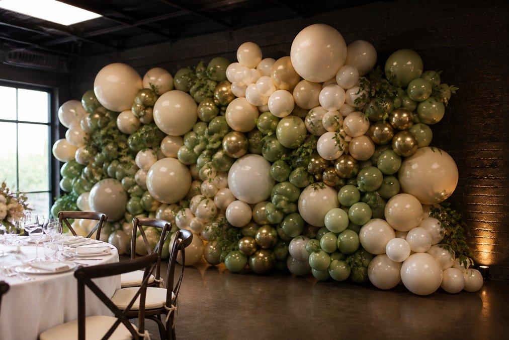 Professional balloon backdrop installation at an event venue showing organic flow with balloon clusters of different sizes, colors, and textures creating a sophisticated aesthetic with natural movement and visual balance for a celebration
